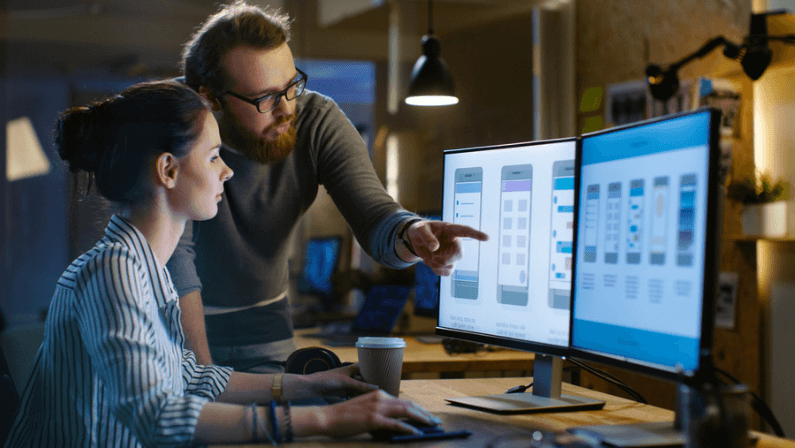 Female UX Architect Has Discussion with Male Design Engineer, They Work on Mobile Application Late at Night. In the Background Wall with Project Sticky Notes and Other Studio Employees